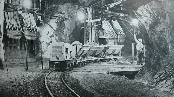 Photographie en noir et blanc d'une galerie avec des ouvriers, un train de wagonnets chargés de roches et l'installation de remplissage en arrière-plan