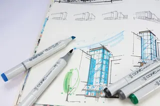 Ein Arbeitsplatz, auf dem eine detaillierte Skizze eines Gebäudes liegt. Mehrere bunte Stifte sind um die Skizze herum verteilt