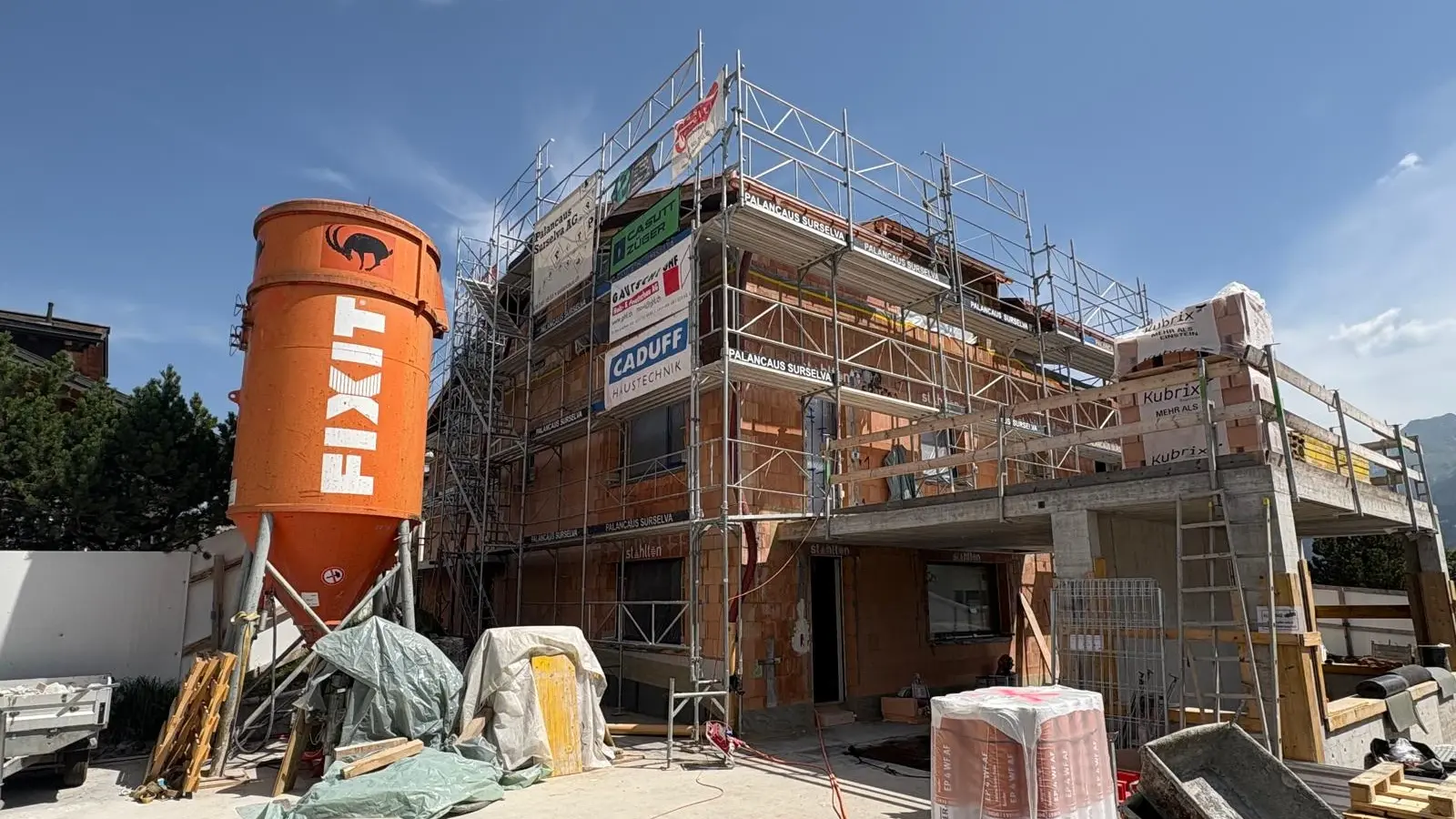 Frontalansicht des eingerüsteten Rohbaus eines MFHs mit links einem orangenen Fixit Silo