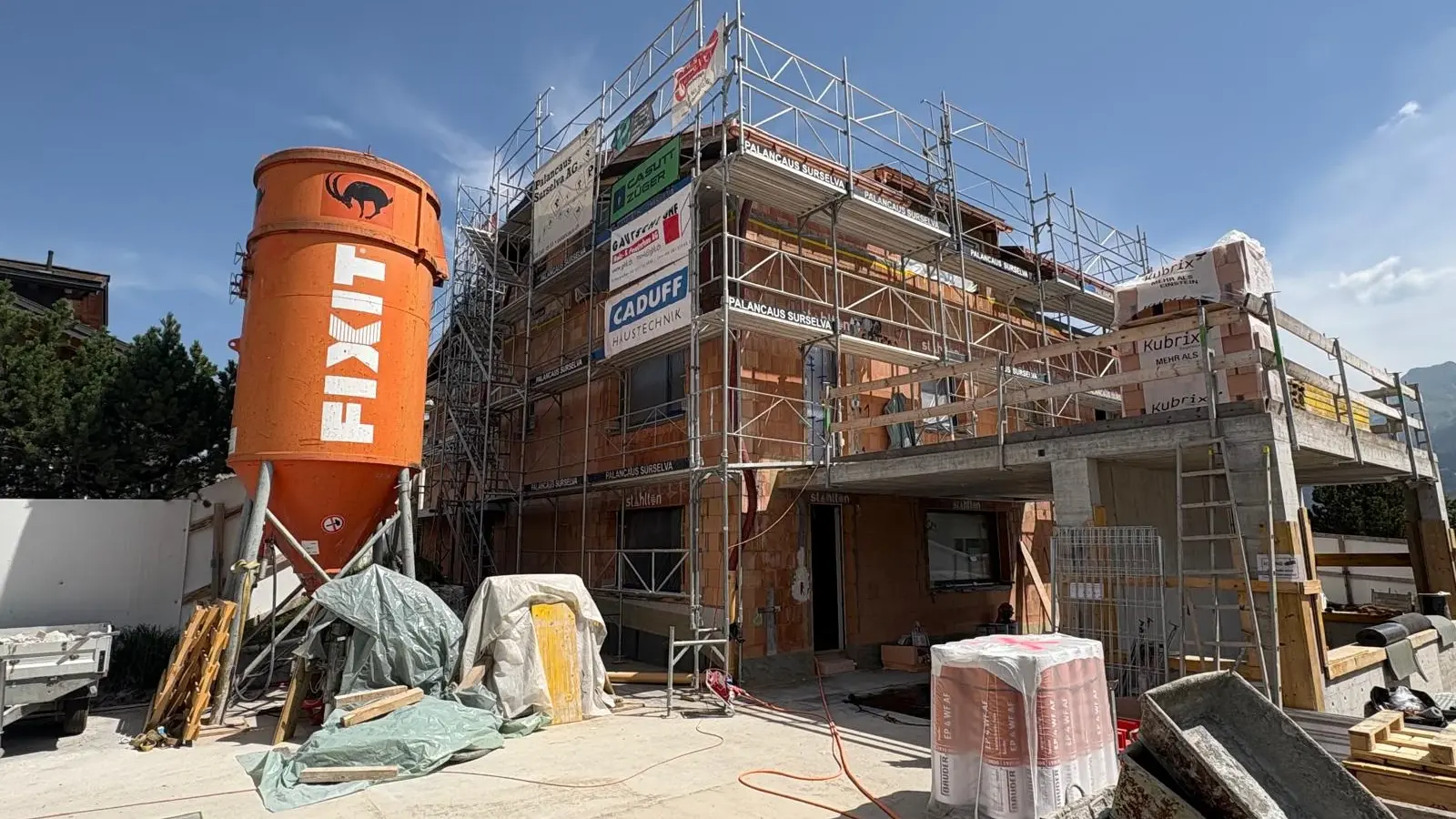 Frontalansicht des eingerüsteten Rohbaus eines MFHs mit links einem orangenen Fixit Silo
