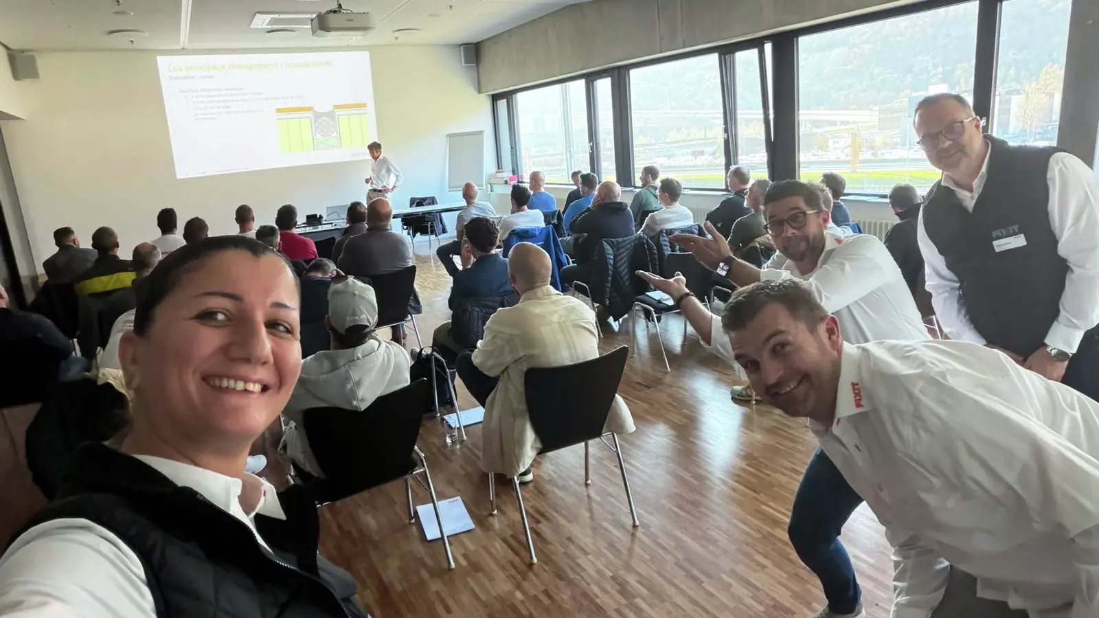 Selfie von Rana Mehaidli mit Samuel Blösch, Kevin Maxwell im Hintergrund die Teilnehmenden sowie Alain Nicaty bei seinem Vortrag