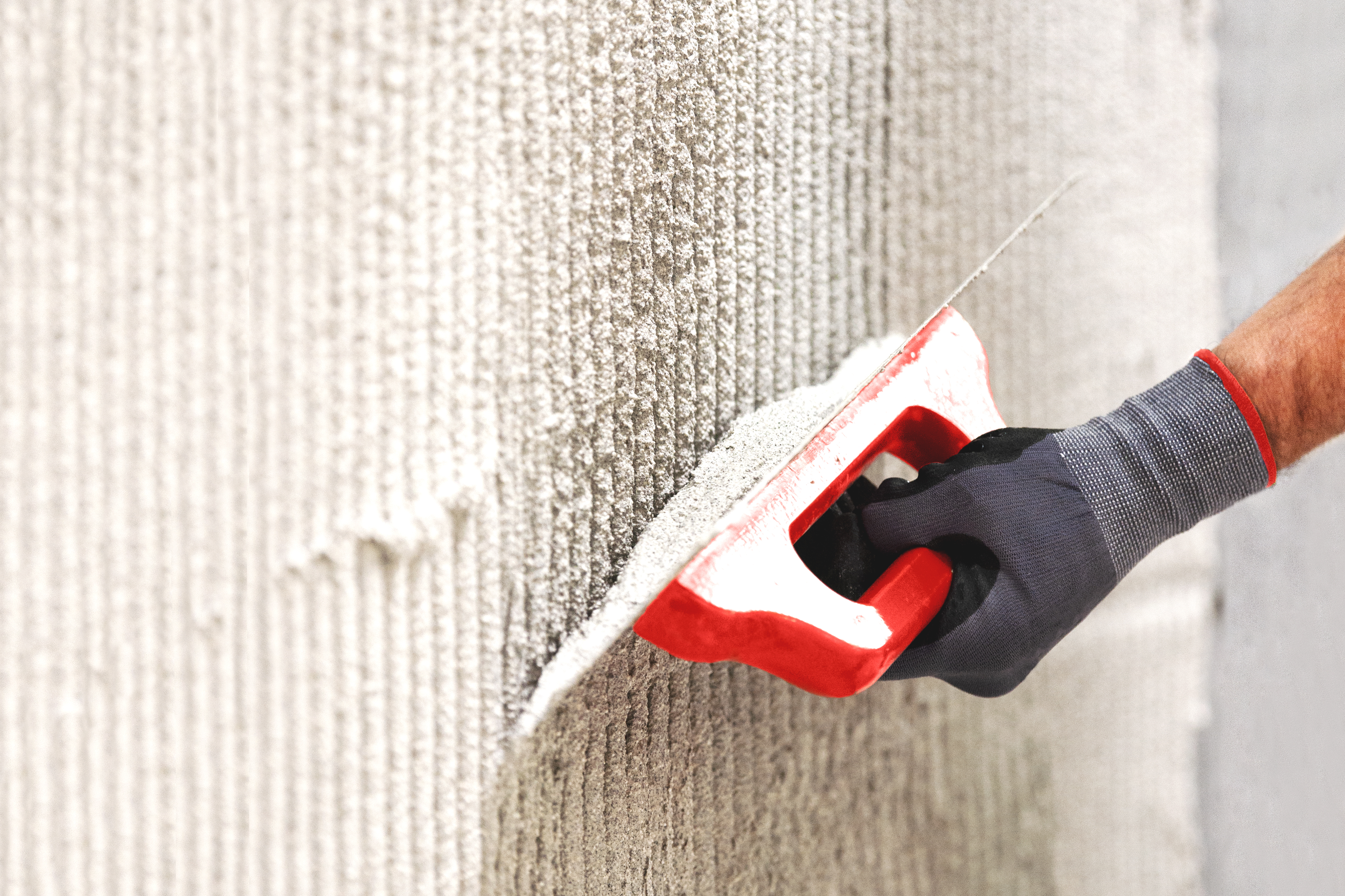 Une main tient une truelle dentée rouge en enlevant du plâtre sur un mur
