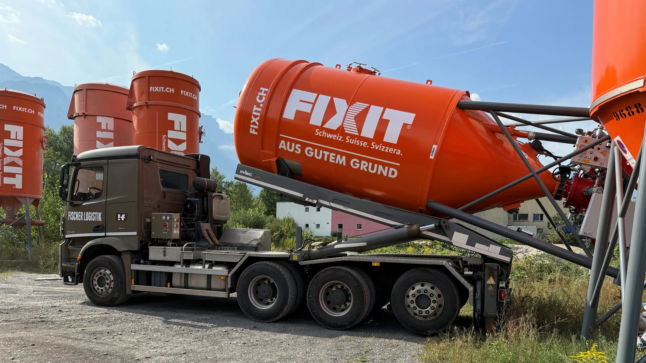 LKW mit orangenem Fixit Clever Drucksilo stellt das Silo auf eine Wiese, weitere Silos, blauer Himmel und Berge im Hintergrund