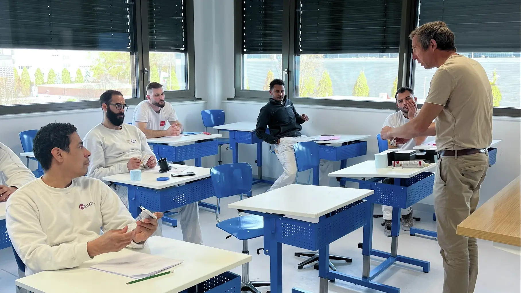 Quatre participants sont assis sur les bancs bleus et blancs et Alain Nicaty explique