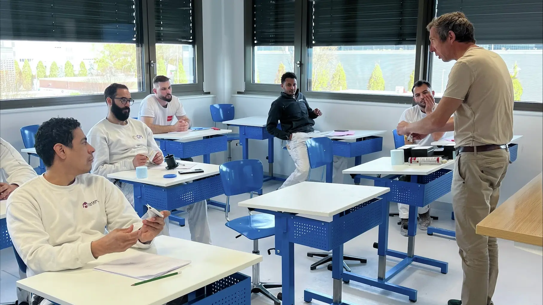 Quatre participants sont assis sur les bancs bleus et blancs et Alain Nicaty explique