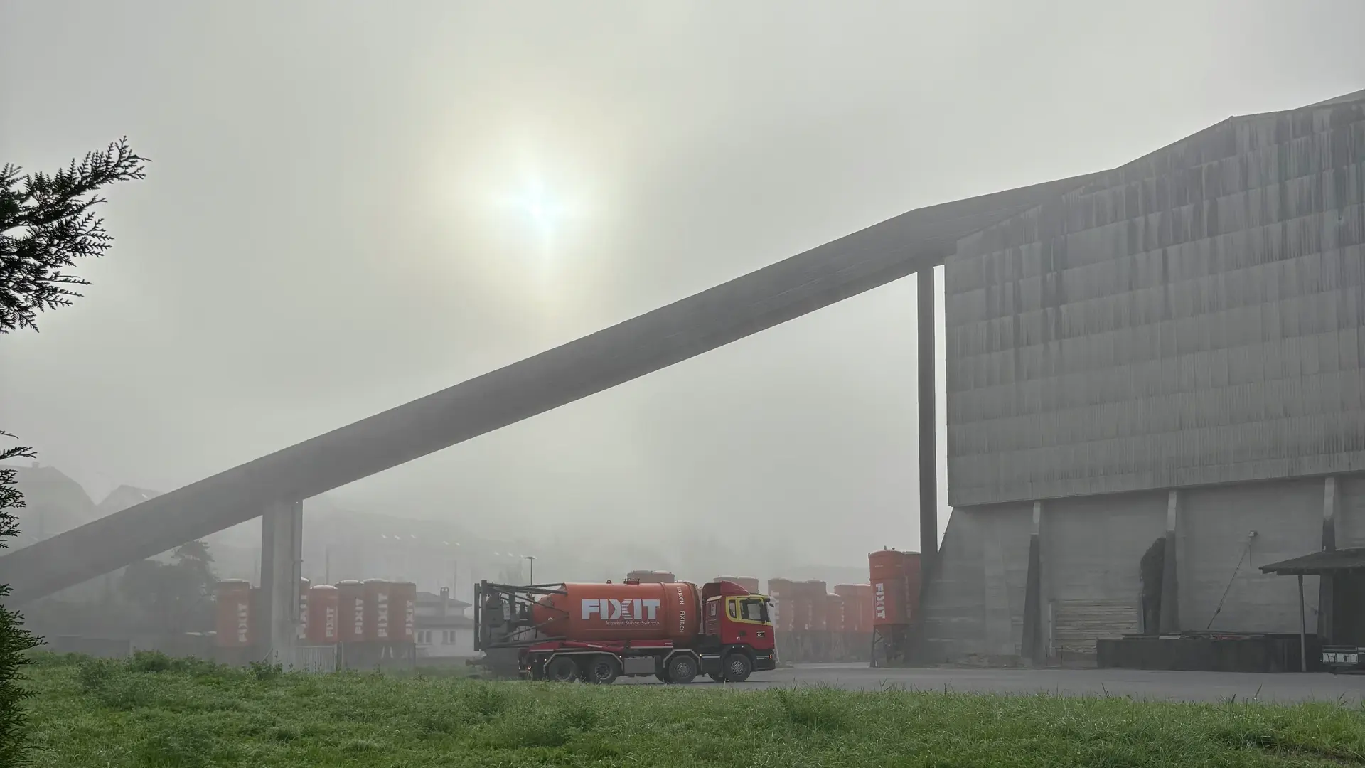 LKW mit orangenem Fixit Silo vor der Steinhalle in Holderbank, die Sonne ist im Nebel verschwunden
