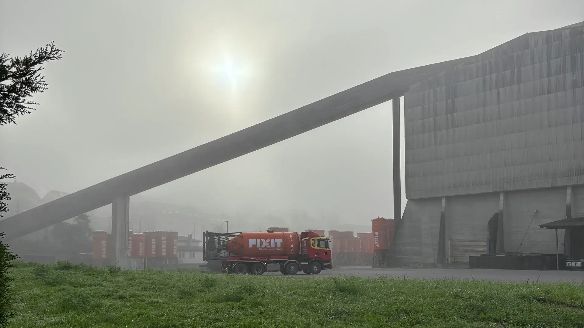 LKW mit orangenem Fixit Silo vor der Steinhalle in Holderbank, die Sonne ist im Nebel verschwunden
