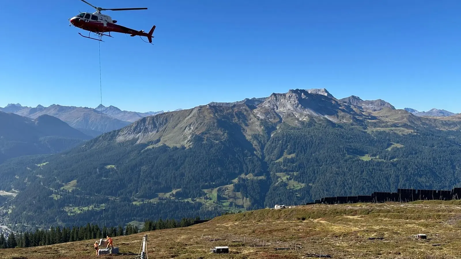 Helikopter lädt eine Palette mit Fixit 532 Ankermörtel auf dem alpinen Installationsgebiet für das Solarkraftwerk Madrisa Solar ab, es sind Bauarbeiter im Vordergrund zu sehen und das Gebirgspanorama im Hintergrund
