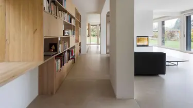 Salon moderne avec une grande étagère en bois pour les livres sur la gauche, un canapé noir, des murs et un plafond en plâtre.