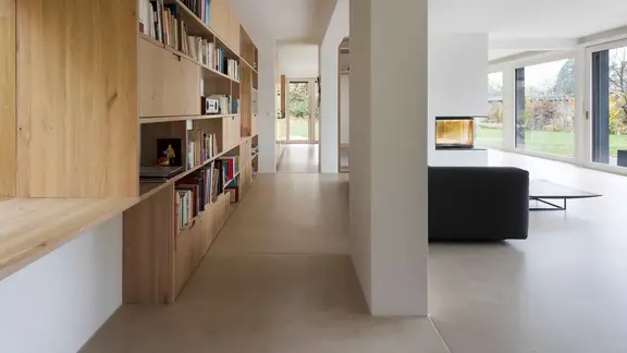 Salon moderne avec une grande étagère en bois pour les livres sur la gauche, un canapé noir, des murs et un plafond en plâtre.