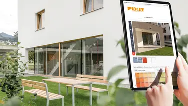 A droite, au premier plan, on voit un plateau avec le configurateur de façade Fixit et les mains de l'utilisateur, à l'arrière-plan, une maison individuelle moderne.