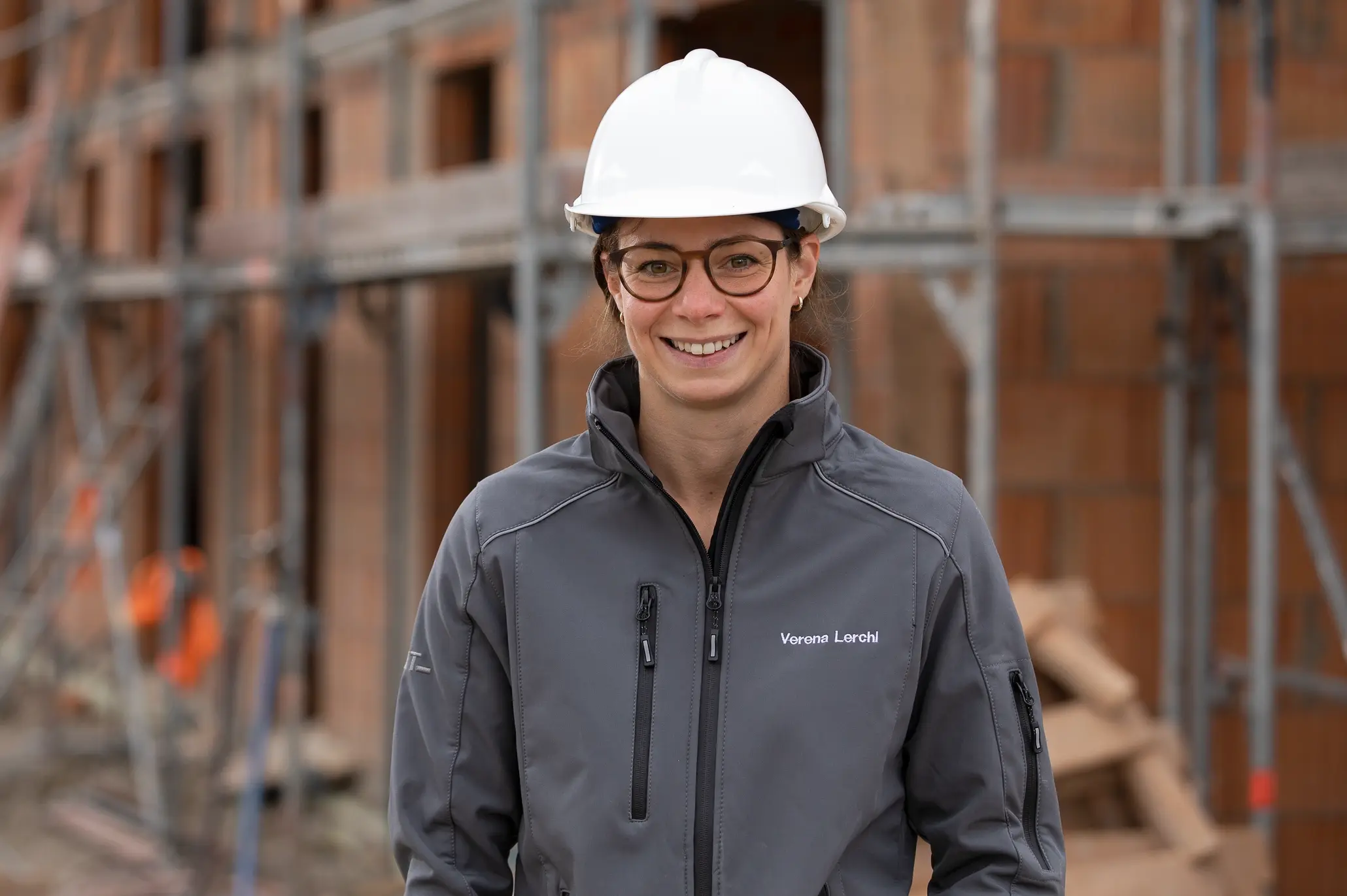 Portrait d'une ouvrière du bâtiment portant un casque de chantier blanc et une veste grise devant un mur de briques Gros œuvre avec échafaudage