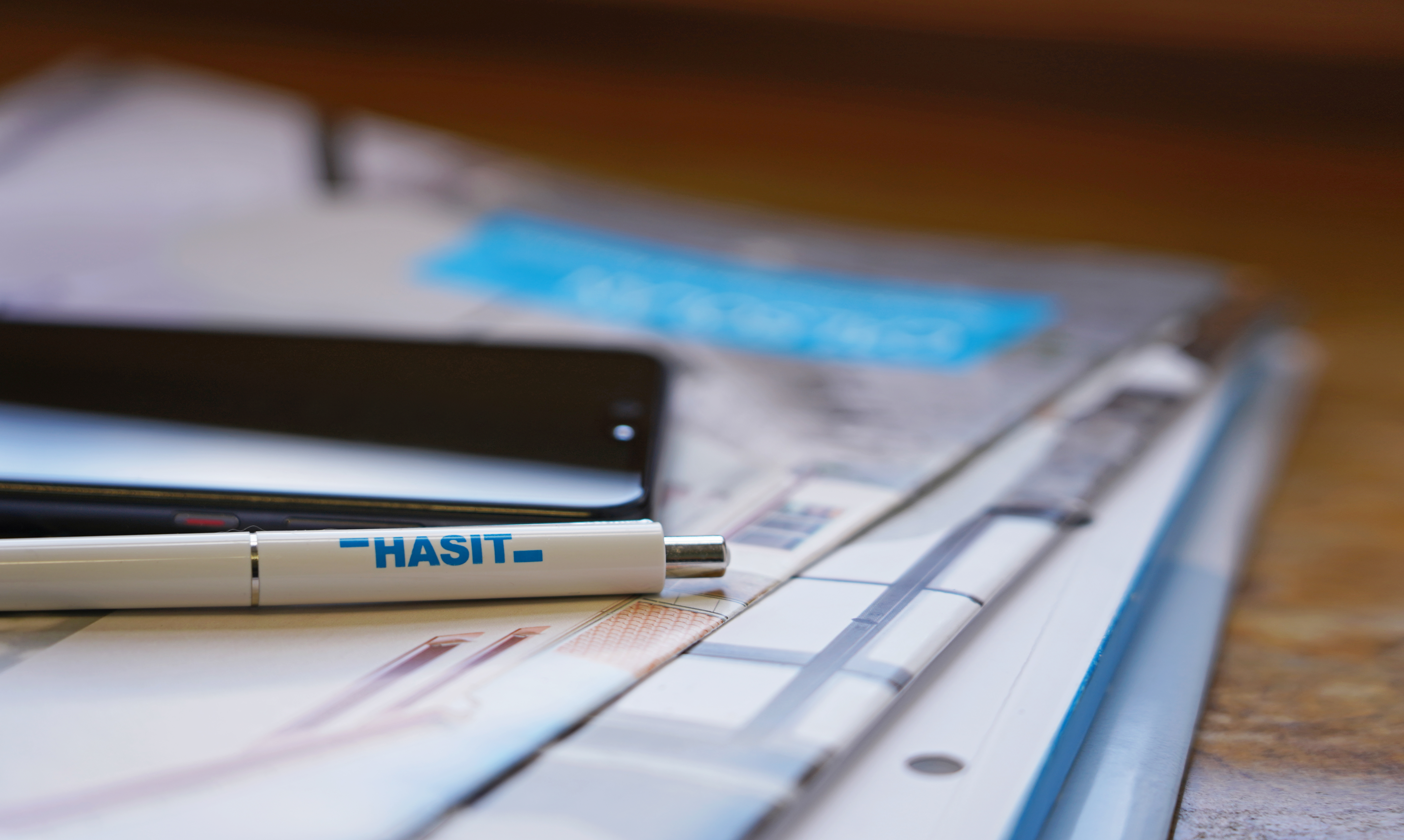 Broschüren und Flyer von HASIT gestapelt auf einem Schreibtisch, zusammen mit einem Handy und einem Kugelschreiber.