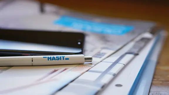 Broschüren und Flyer von HASIT gestapelt auf einem Schreibtisch, zusammen mit einem Handy und einem Kugelschreiber.