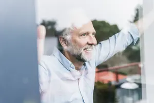 Älterer Mann schaut zufrieden aus dem Fenster und genießt die Vorteile seines energieeffizient gedämmten Hauses.