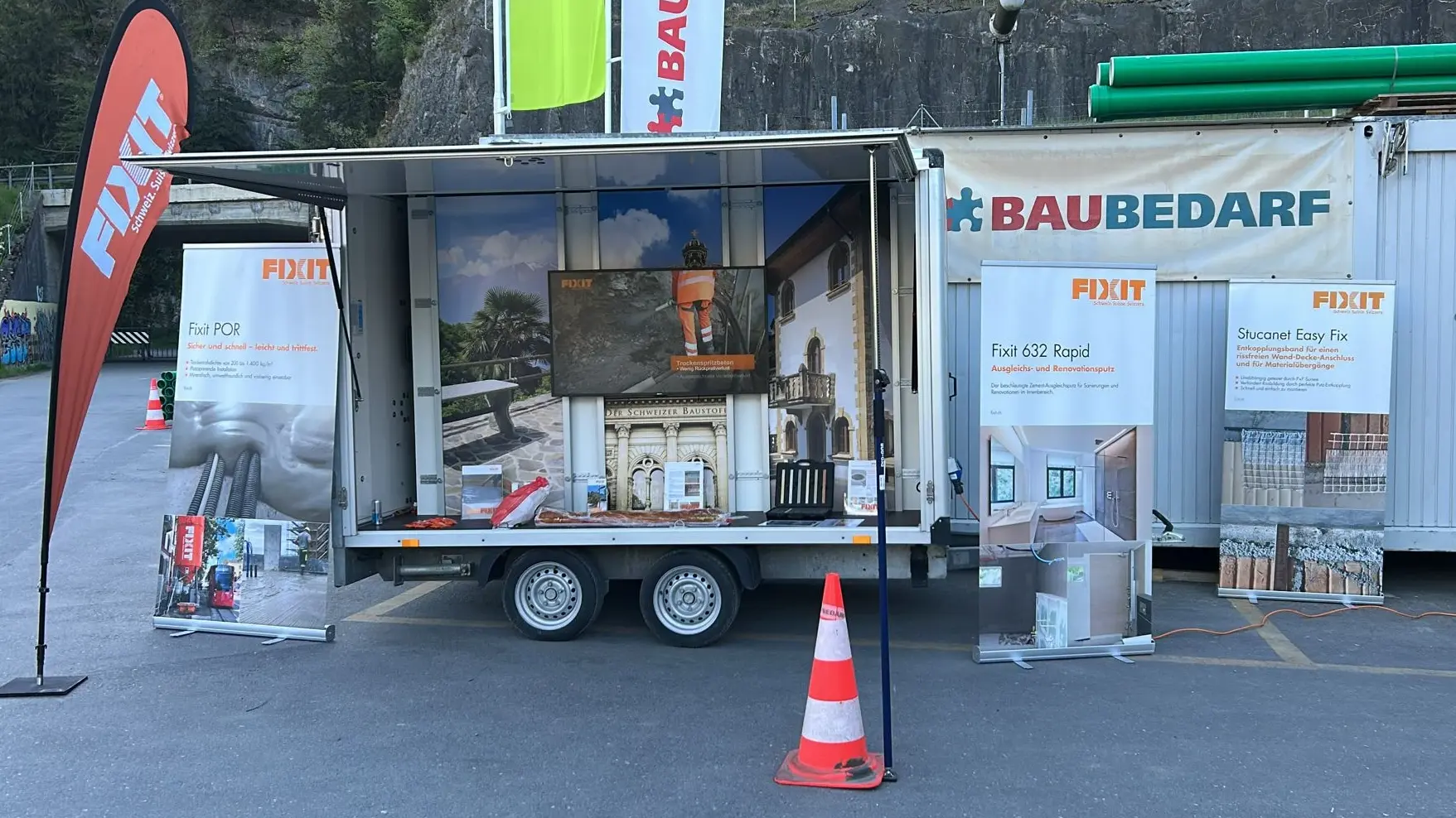 Remorque d'exposition Fixit dépliée, équipée d'un écran, d'un drapeau de plage orange, de bannières enroulables, d'un cône de signalisation et du logo de Baubedarf