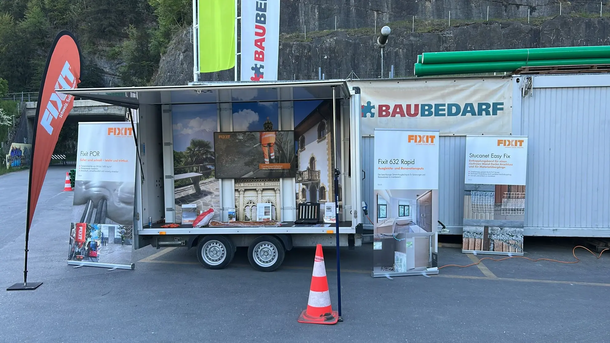 Remorque d'exposition Fixit dépliée, équipée d'un écran, d'un drapeau de plage orange, de bannières enroulables, d'un cône de signalisation et du logo de Baubedarf