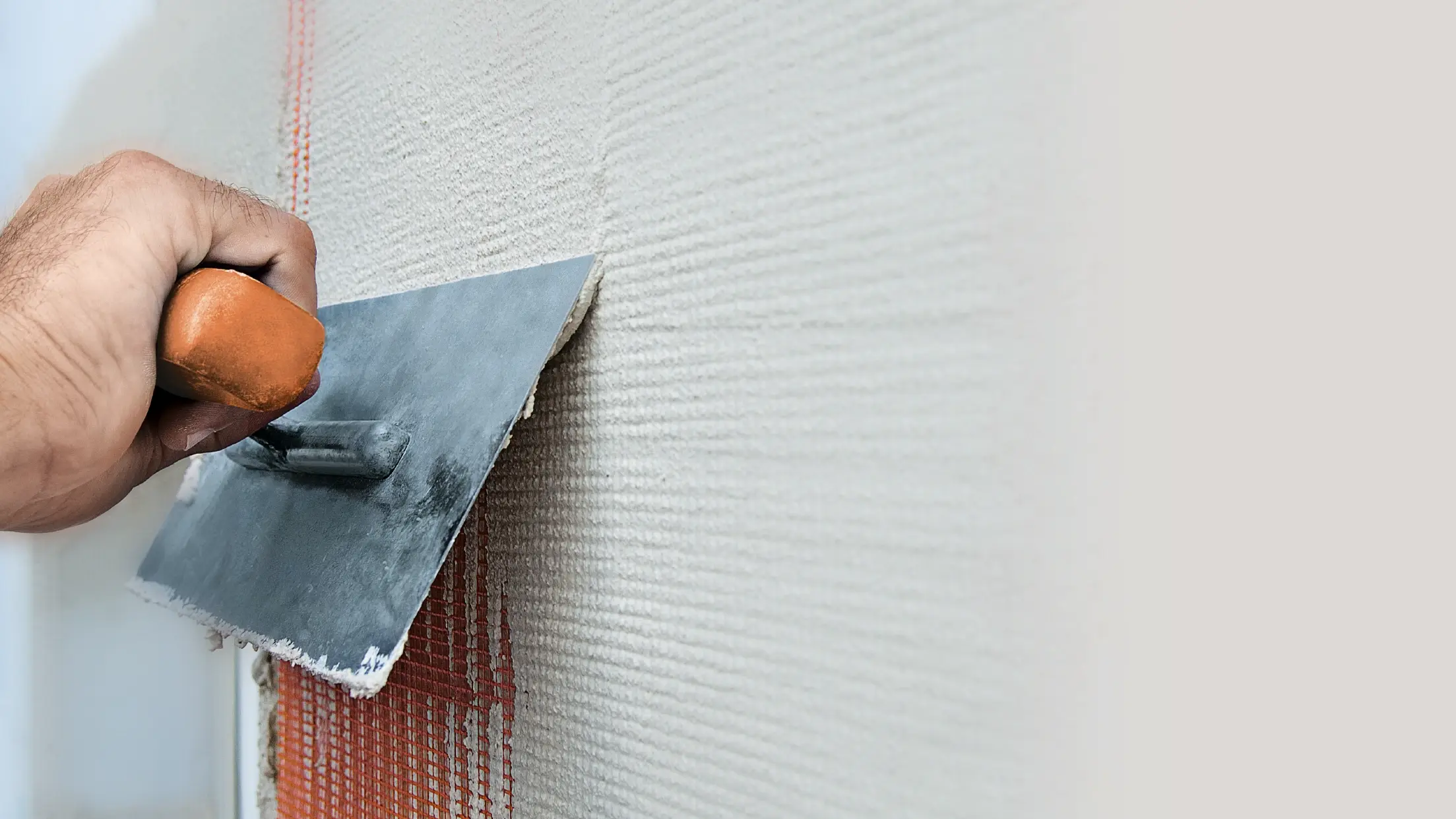 Nahaufnahme einer Wand mit Verputz, eine Hand zieht mit einer Metallkelle Putz über ein Armierungsnetz