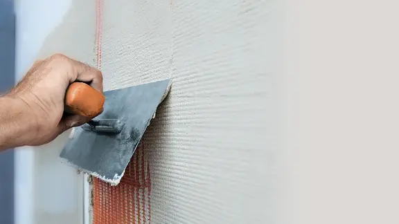 Nahaufnahme einer Wand mit Verputz, eine Hand zieht mit einer Metallkelle Putz über ein Armierungsnetz