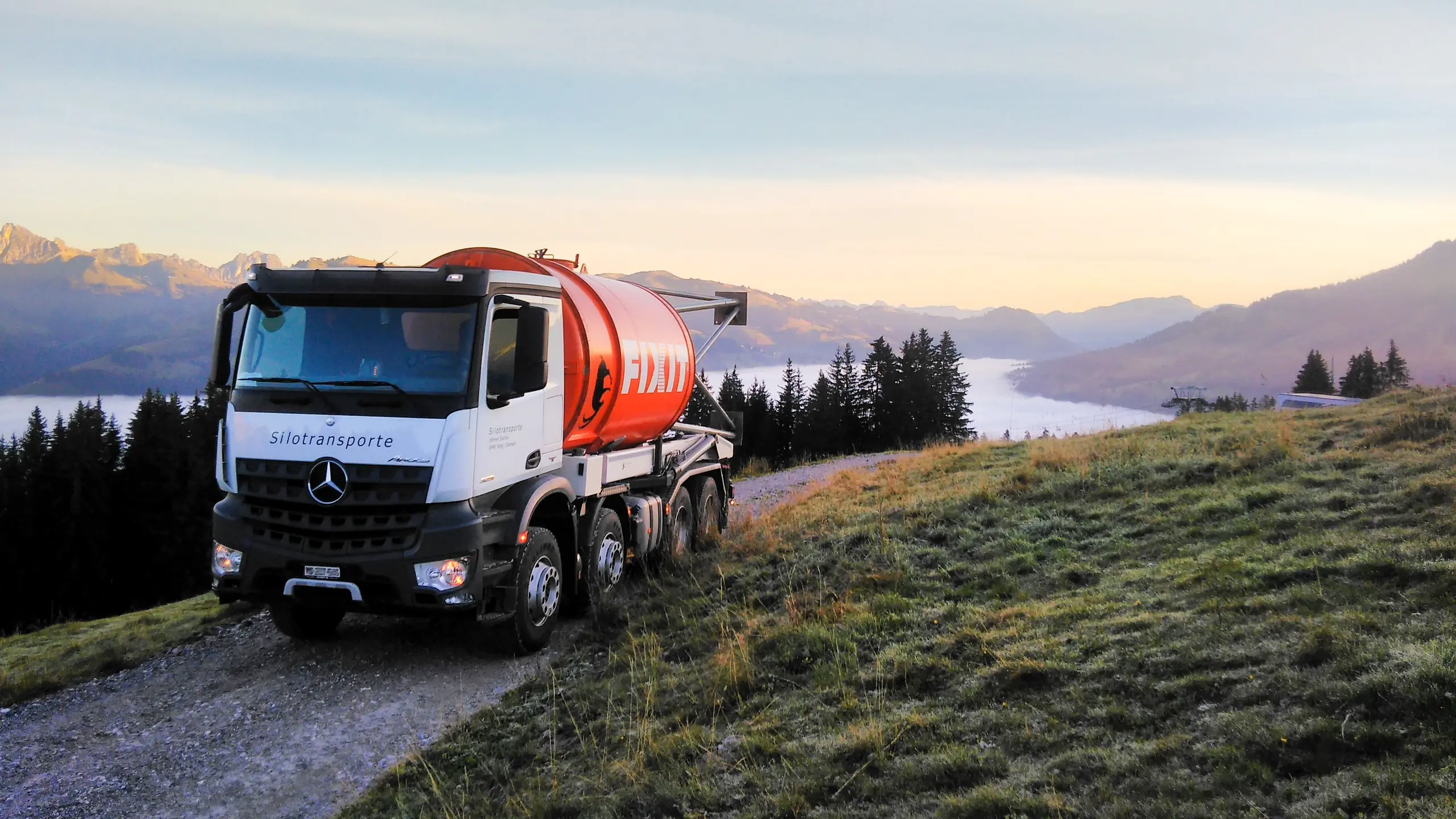 Camion blanc avec un silo Fixit orange sur une route de gravier, à l'aube, avec un panorama montagneux