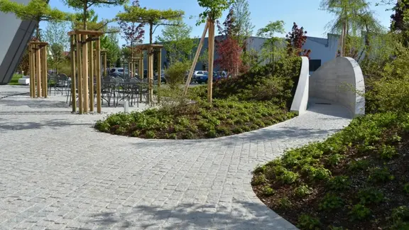 Parvis moderne avec des pavés gris clair et des plates-bandes plantées de vert ainsi que des arbres isolés