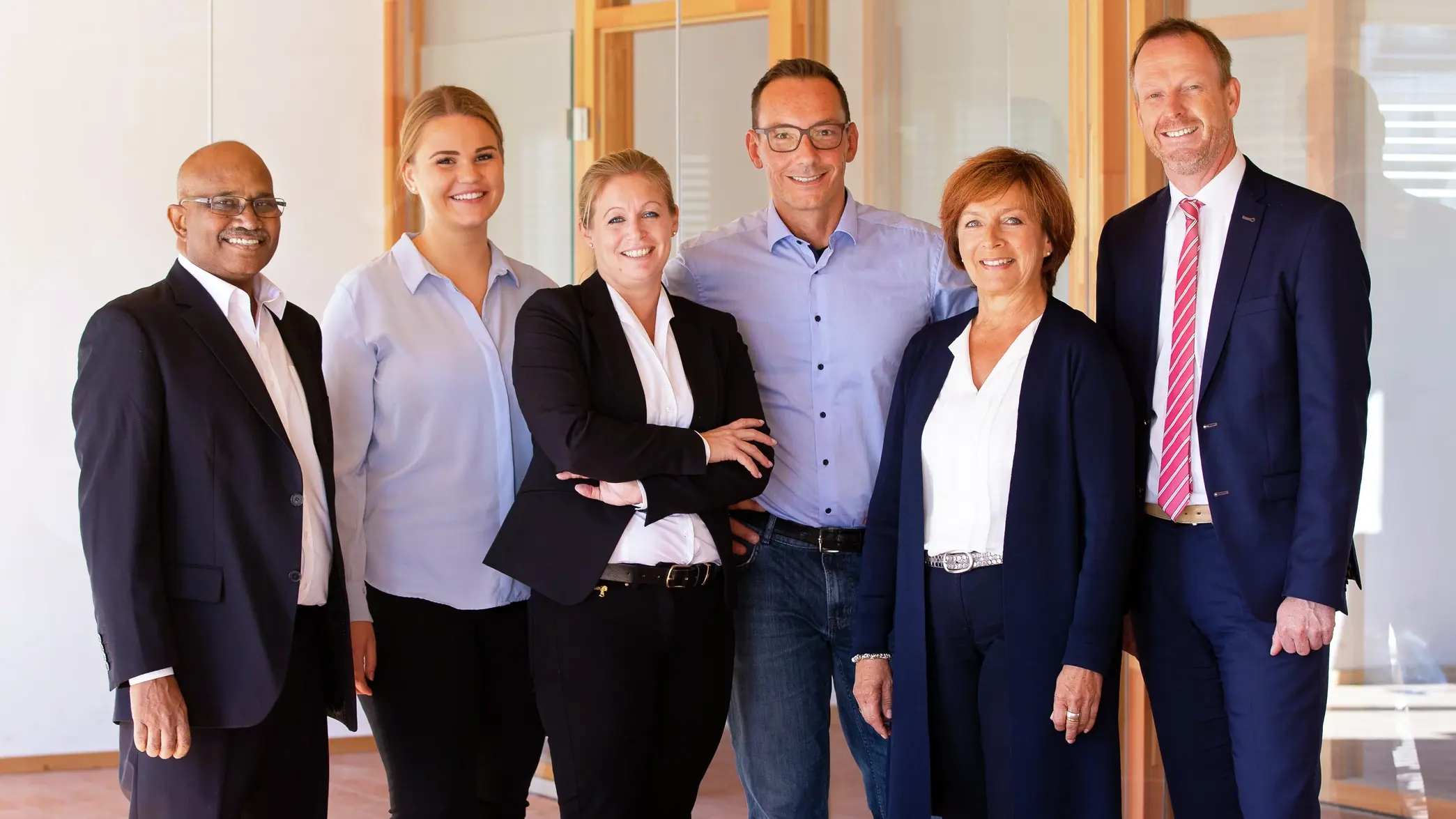 Photo de groupe avec six personnes, dont deux femmes, toutes vêtues d'une chemise et d'un costume, devant une baie vitrée en bois