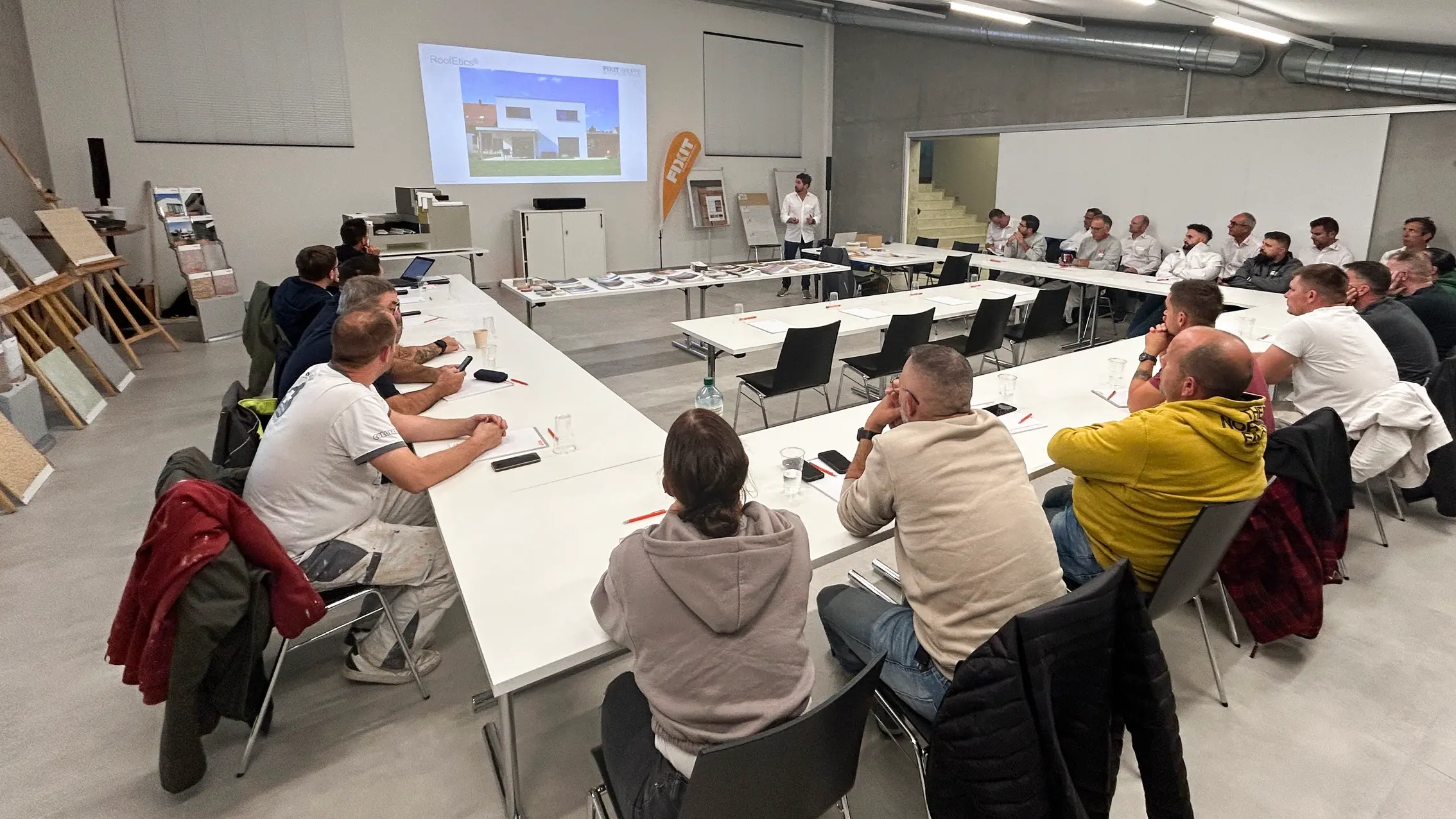 Les participants sont assis à des tables disposées en U et écoutent la conférence.
