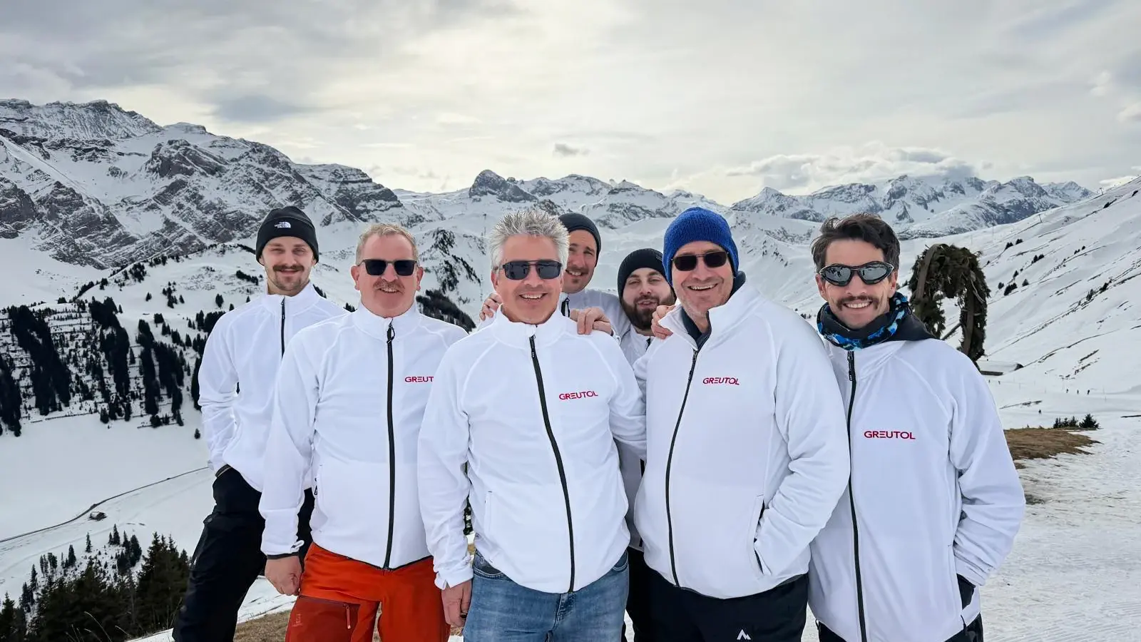 Sieben Greutol Mitarbeiter mit weissen Greutol-Jacken als Gruppe vor dem Bergpanorama