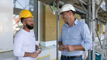 Un transformateur parle à un commercial en chemise bleu clair devant une façade extérieure avec des fenêtres installées et un échafaudage.