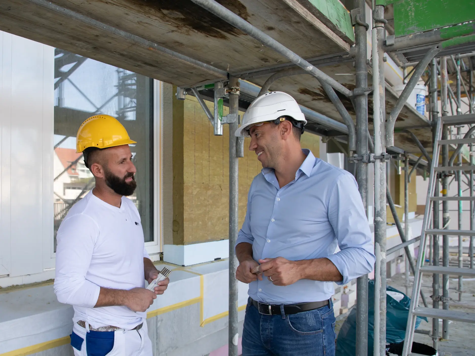 Ein Vertriebsmitarbeiter in hellblauem Hemd mit weissem Bauhelm redet auf einem Baugerüst mit einem Verarbeiter in weisser Kleidung und gelben Bauhelm