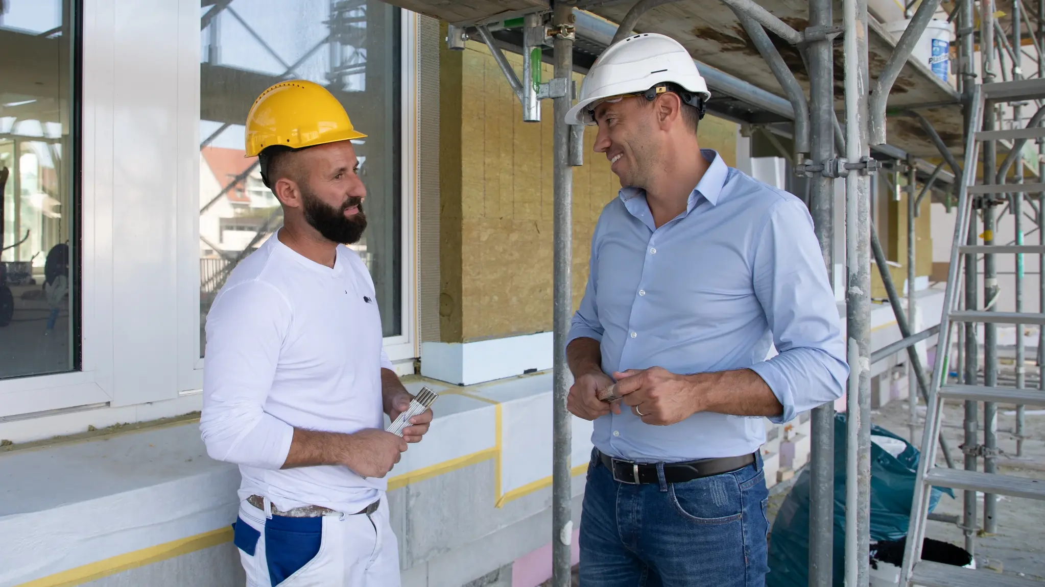 Un artisan discute avec un commercial en chemise bleu clair devant une façade extérieure dotée de fenêtres intégrées et d’échafaudages