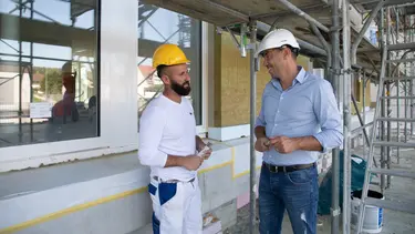 Ein Bauarbeiter spricht mit einem Vertriebsmitarbeiter im Hemd auf einem Baugerüst vor einer Fassade
