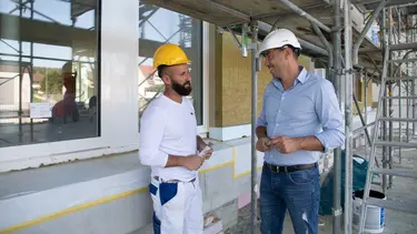 Ein Verarbeiter spricht mit einem Vertriebsmitarbeiter in hellblauem Hemd vor einer Aussenfassade mit eingebauten Fenstern und Baugerüst