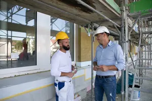 Ein Bauarbeiter spricht mit einem Vertriebsmitarbeiter im Hemd auf einem Baugerüst vor einer Fassade