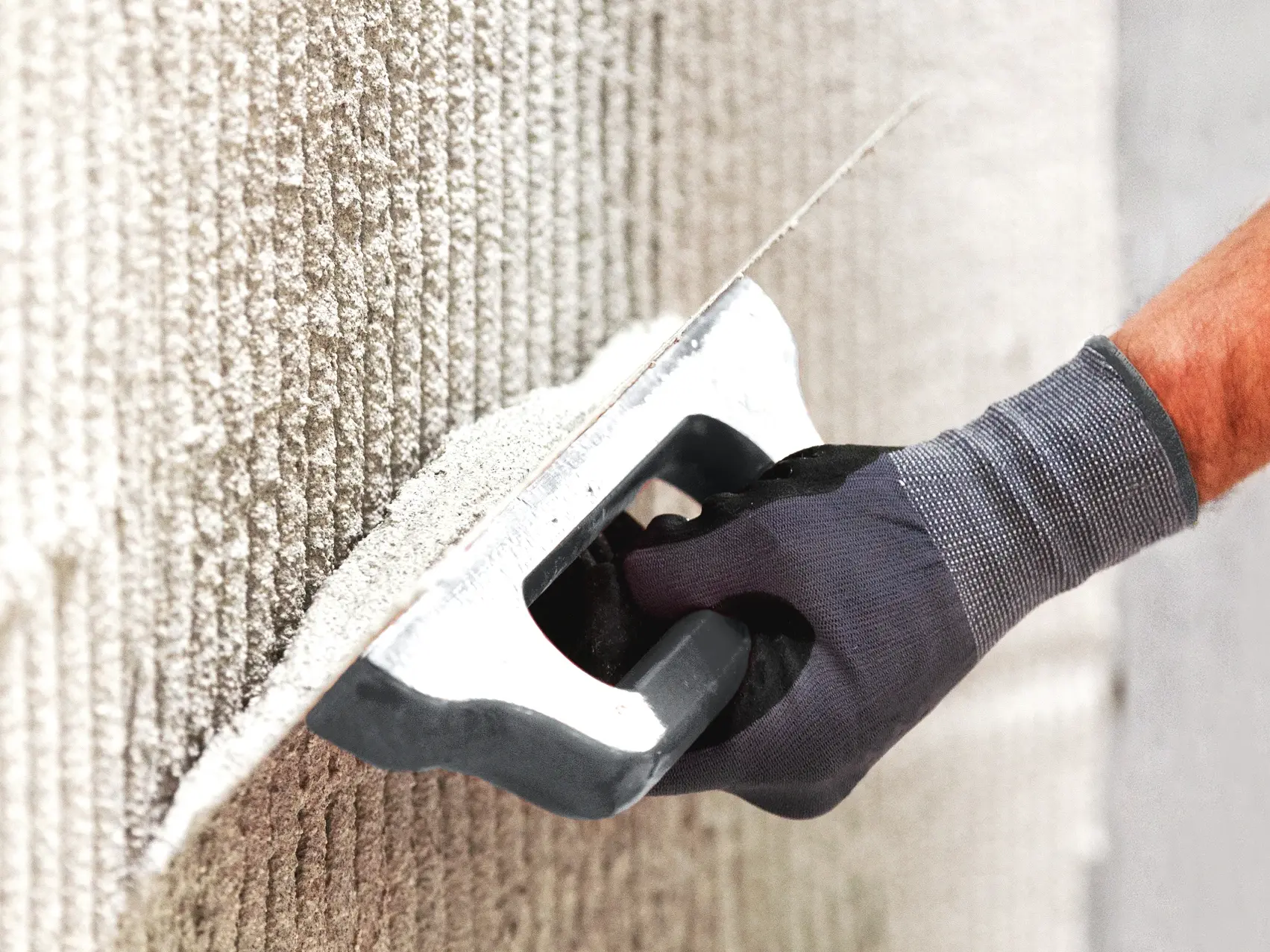 Verputz wird mit einer Zahntraufel von einer Hand in einem grauen Arbeitshandschuh aufgezogen.